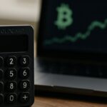 Uitgeschakelde hardwarewallet op een bureau met op de achtergrond een laptop met Bitcoin-koersgrafiek