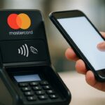Betaalterminal met Mastercard-logo waarbij iemand met een smartphone contactloos betaalt