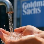 Helder water stroomt uit een kraan in gevouwen handen, met op de achtergrond het logo van Goldman Sachs op een gevel.