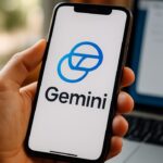 Een hand houdt een smartphone vast met de Google Gemini-app, op een cafétafel naast een laptop