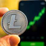Fysieke zilveren Litecoin-munt met het L-logo, op de achtergrond een telefoon met een stijgende groene grafiek