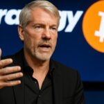 Michael Saylor spreekt bij een persmoment, met op de achtergrond het Strategy-logo en een oranje bitcoin-symbool