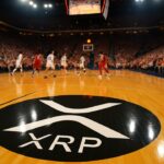 XRP-logo prominent op de houten basketbalvloer van een volle Amerikaanse sportarena tijdens een wedstrijd
