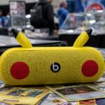 Gele Beats Pill-speaker in Pikachu-stijl met Beats-logo op een stapel Pokémonkaarten op een verzamelbeurs