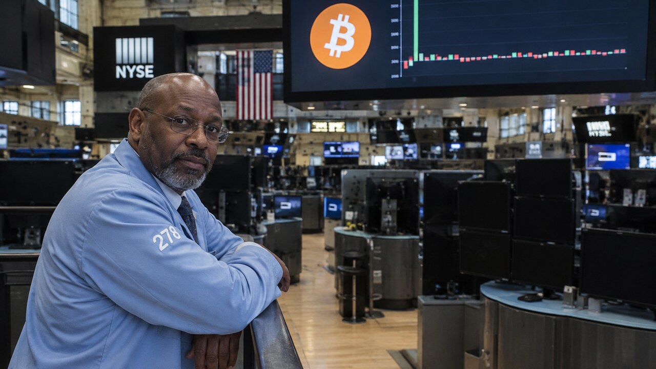 Oudere beurshandelaar leunt op een reling op een bijna lege beursvloer onder een scherm met het Bitcoin-symbool en een afgevlakte grafiek van fondsstromen