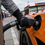 Monteur tankt een oranje raceauto met een groot Bitcoin-logo bij een pitstop, terwijl de brandstofmeter nog maar een kwart vol staat
