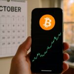 Hand met smartphone die een stijgende Bitcoin-grafiek toont voor een kalender op de maand oktober, met herfstbladeren buiten