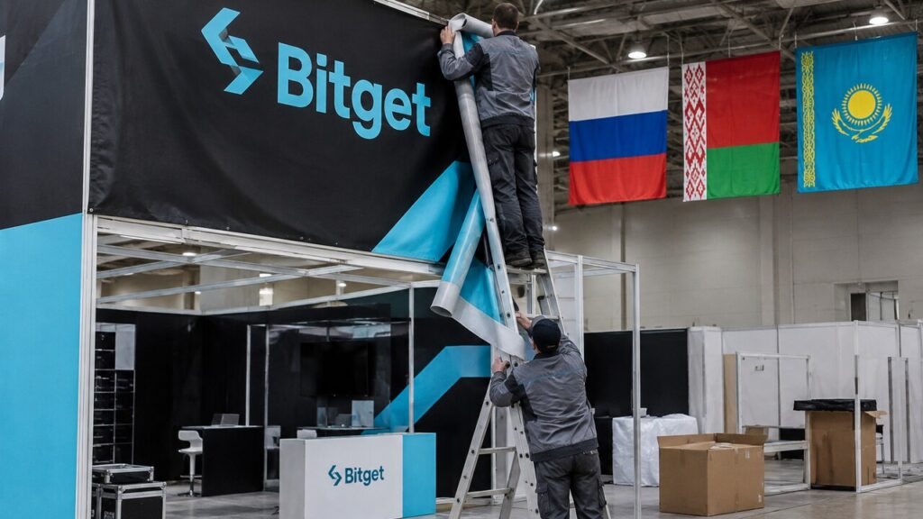 Medewerkers halen een groot Bitget-spandoek neer op een beursstand, met de vlaggen van Rusland, Belarus en Kazachstan op de achtergrond
