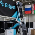 Medewerkers halen een groot Bitget-spandoek neer op een beursstand, met de vlaggen van Rusland, Belarus en Kazachstan op de achtergrond