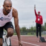 Sprinter in startblokken op een atletiekbaan met het Canton-logo op zijn shirt, klaar voor de start