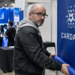 Man rolt een banier met het Cardano-logo op bij een lege beursstand, terwijl op de achtergrond een drukke stand met het Sui-logo staat
