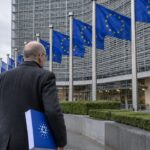Man loopt met een document met het Cardano-logo naar het Berlaymont-gebouw van de Europese Commissie in Brussel, met EU-vlaggen ervoor