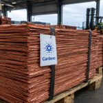 Stapels koperkathodes op pallets in een havenloods, met een label met het Cardano-logo