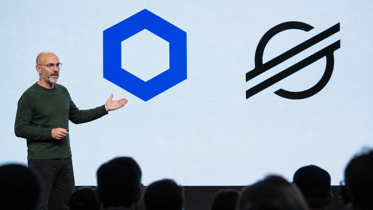 Spreker op een podium voor een groot scherm met de logo's van Chainlink en Stellar tijdens een ontwikkelaarsconferentie