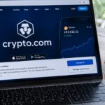 Laptop op een keukentafel met de Crypto.com-website en een cookiemelding in beeld, naast een koekje op een bordje
