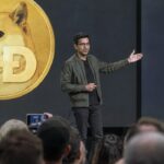 Presentator op een podium van een cryptoconferentie voor een groot scherm met het Dogecoin-logo, terwijl het publiek toekijkt