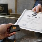 Een hand schuift een Bitcoin hardware wallet over een marmeren balie in ruil voor een aandelenbewijs met het Grayscale-logo