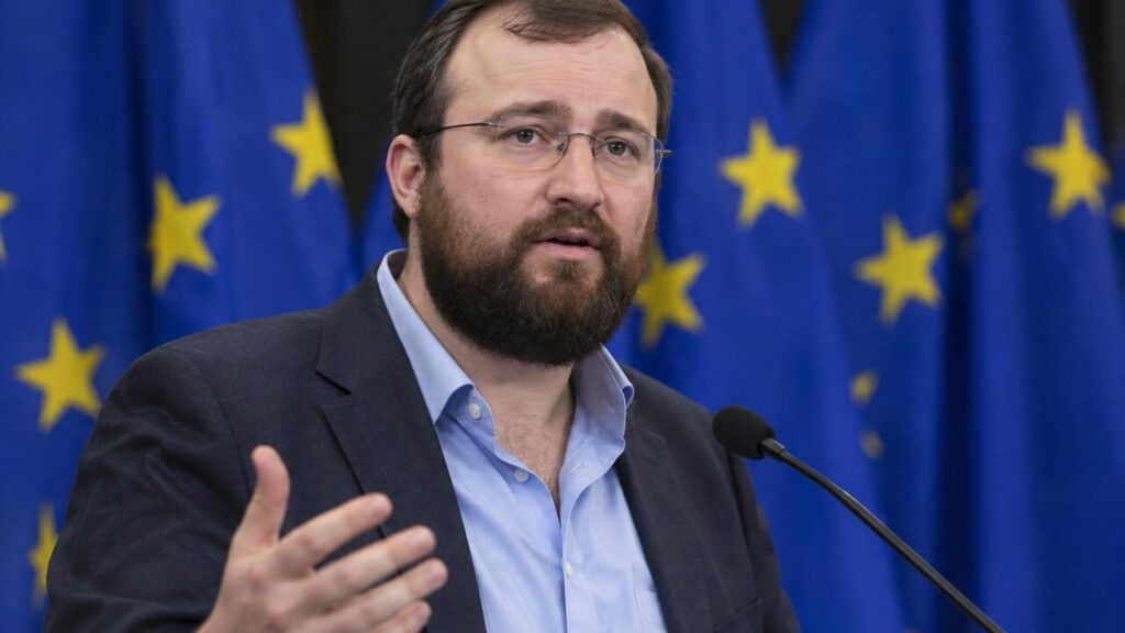 Cardano-oprichter Charles Hoskinson spreekt op een podium met Europese vlaggen op de achtergrond