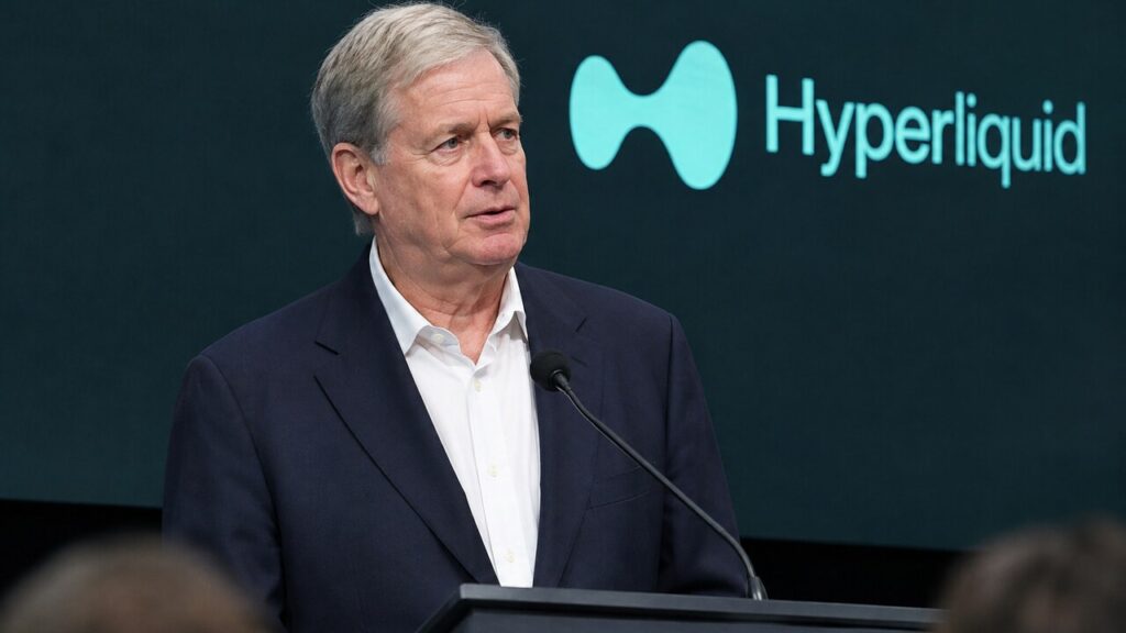 Belegger Stanley Druckenmiller op een conferentiepodium met het Hyperliquid-logo op het scherm achter hem