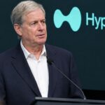 Belegger Stanley Druckenmiller op een conferentiepodium met het Hyperliquid-logo op het scherm achter hem