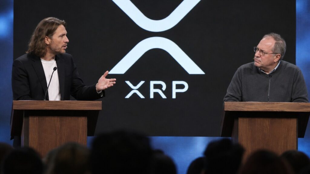 Twee sprekers in debat op een podium van een blockchainconferentie voor een groot scherm met het XRP-logo