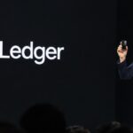 Presentator op het podium van Ledger Op3n in Parijs houdt de Ledger Atom omhoog, met het Ledger-logo op het scherm
