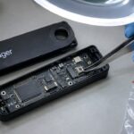 Opengemaakte Ledger hardware wallet op een werkbank, waarbij een pincet onder een vergrootlamp een extra onderdeel in het apparaat aanwijst