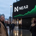 Groot reclamescherm op een plein met het NEAR-logo en een stijgende groene koersgrafiek, met voorbijgangers die omhoog kijken