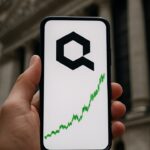 Hand houdt smartphone met stijgende Quant-koersgrafiek voor een bankgebouw in New York
