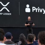 Podium van een ontwikkelaarsconferentie met een groot scherm met de logo's van XRP Ledger en Privy en een spreker voor publiek