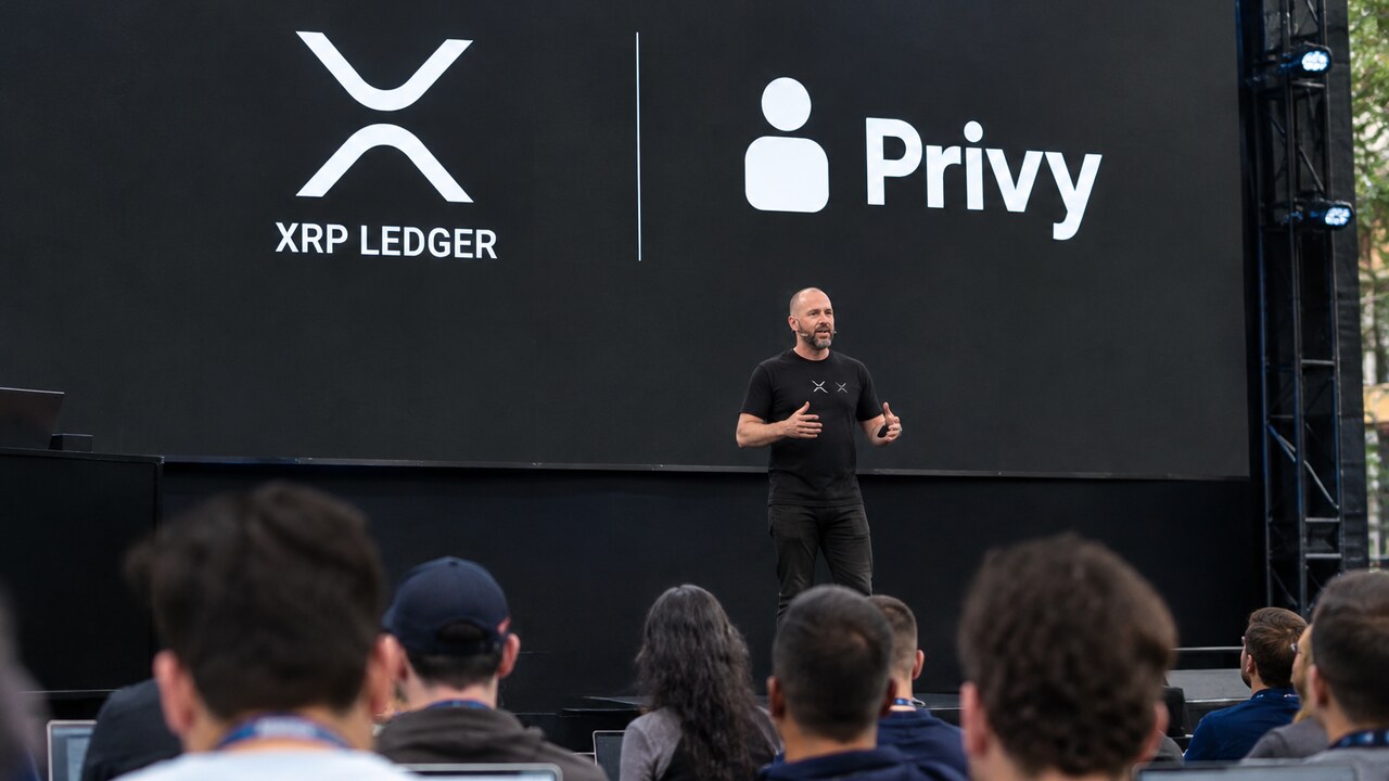 Podium van een ontwikkelaarsconferentie met een groot scherm met de logo's van XRP Ledger en Privy en een spreker voor publiek