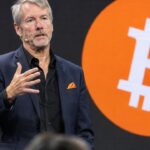 Michael Saylor spreekt op een podium met een groot oranje Bitcoin-logo op het scherm achter hem