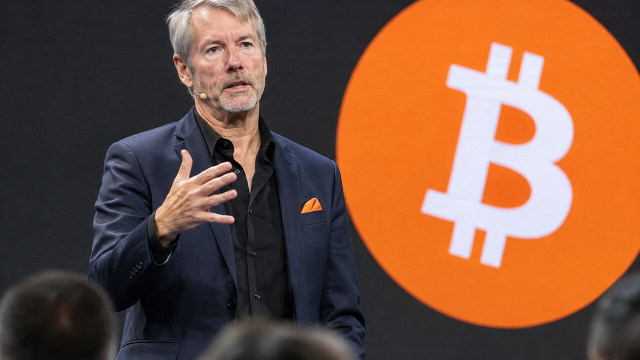 Michael Saylor spreekt op een podium met een groot oranje Bitcoin-logo op het scherm achter hem