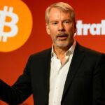 Michael Saylor spreekt op een podium met het Bitcoin-symbool en het Strategy-logo op een groot scherm achter hem