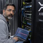 Beveiligingsengineer knielt in een datacenter naast een serverrack met een groot XRP-logo en controleert code op een laptop