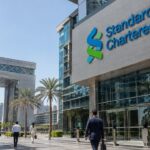 Gevel van een Standard Chartered-gebouw in het financiële centrum van Dubai met een groot bankslogo, palmbomen en voorbijgangers in de middagzon