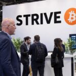 Stand van Strive op een Bitcoin-conferentie met een groot Strive-logo en Bitcoin-symbool, met bezoekers die erlangs lopen
