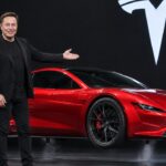 Elon Musk op een podium naast een rode Tesla Roadster die net boven de vloer zweeft, met het Tesla-logo op de achtergrond