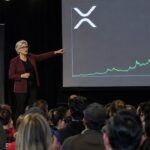 Spreker op een cryptobijeenkomst wijst naar een groot scherm met het XRP-logo en een steil stijgende groene koersgrafiek, met publiek op de voorgrond