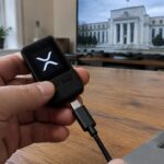 Hand haalt een hardware wallet met het XRP-logo van een laptop, met op de achtergrond een televisie met het gebouw van de Federal Reserve